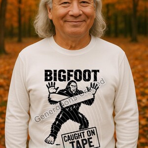 May include: White long-sleeve shirt with a Bigfoot graphic. The design features the word "BIGFOOT" above an illustration of the creature, with the text "CAUGHT ON TAPE" below. The shirt is a comfortable, casual style.