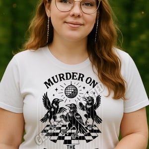 May include: White t-shirt with a black graphic featuring three crows, a disco ball, and the text "MURDER ON THE DANCE FLOOR." The design is set against a checkered floor pattern. The text "GENERATIONS CRAFTED" is also visible.