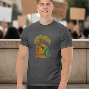 May include: Dark gray t-shirt featuring a graphic of a weeping Statue of Liberty leaning against a tombstone under a tree. The tombstone reads "Freedom of the United States" and "Died 2023".