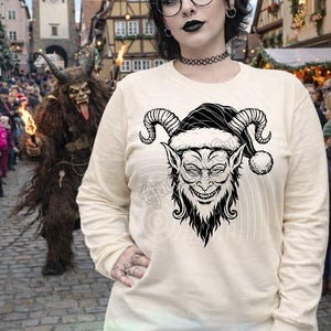 May include: Cream-colored long-sleeve shirt featuring a black and white illustration of a horned, grinning creature wearing a Santa hat. The text "Generations Crafted" is visible at the bottom of the shirt. The background shows a European town.