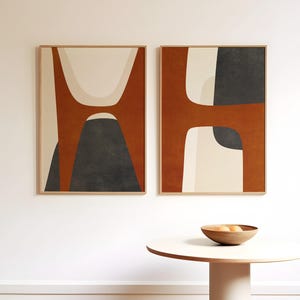 May include: Two framed abstract art prints. Each print features a design with rust, cream, and dark grey shapes. The frames are made of light-coloured wood. A small, round table with a wooden bowl on top is in the foreground.