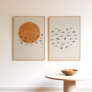 May include: Two framed art prints. One features an orange sun with black bird silhouettes. The other print shows a heart shape formed by black bird silhouettes. Both prints have a beige background and are in light brown frames.
