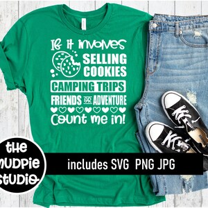 May include: Green t-shirt with white text that reads "If it involves selling cookies, camping trips, friends or adventure, count me in!"  The t-shirt is on a white wooden surface with a pair of blue jeans and black sneakers.