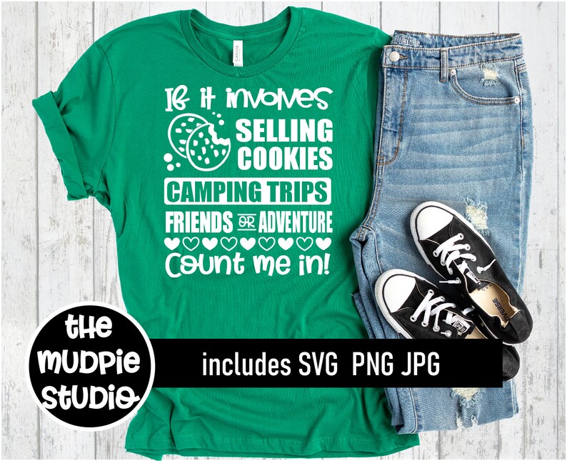 May include: Green t-shirt with white text that reads "If it involves selling cookies, camping trips, friends or adventure, count me in!"  The t-shirt is on a white wooden surface with a pair of blue jeans and black sneakers.