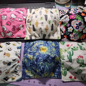 May include: Six square fabric bowl cozies with various patterns. Designs include Hello Kitty, Star Wars characters, sugar skulls, sushi, Van Gogh's "Starry Night", and cacti. The cozies are quilted and appear to be made of cotton.