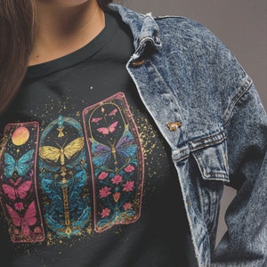 May include: Black t-shirt featuring a graphic of three colourful, ornate panels with butterflies and floral designs. The panels are set against a black background with gold glitter.
