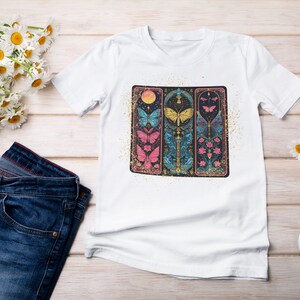 May include: White t-shirt featuring a graphic print of three vertical panels with butterflies, flowers, and a crescent moon. The panels are outlined in gold and have a glittery background.
