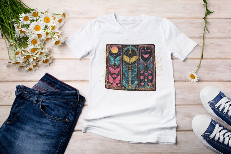 May include: White t-shirt featuring a graphic print of three vertical panels with butterflies, flowers, and a crescent moon. The panels are outlined in gold and have a glittery background.