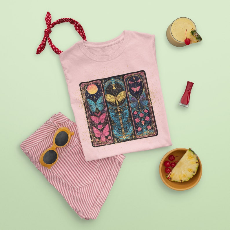 May include: Pink t-shirt featuring a graphic of three colourful butterflies within ornate frames. The butterflies are surrounded by flowers and leaves. The shirt has a distressed look.