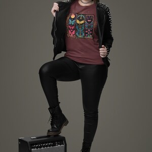 May include: A woman wearing a maroon t-shirt with a colourful butterfly graphic, a black leather jacket, black trousers, and black boots. She is standing with one foot on a black guitar amplifier.
