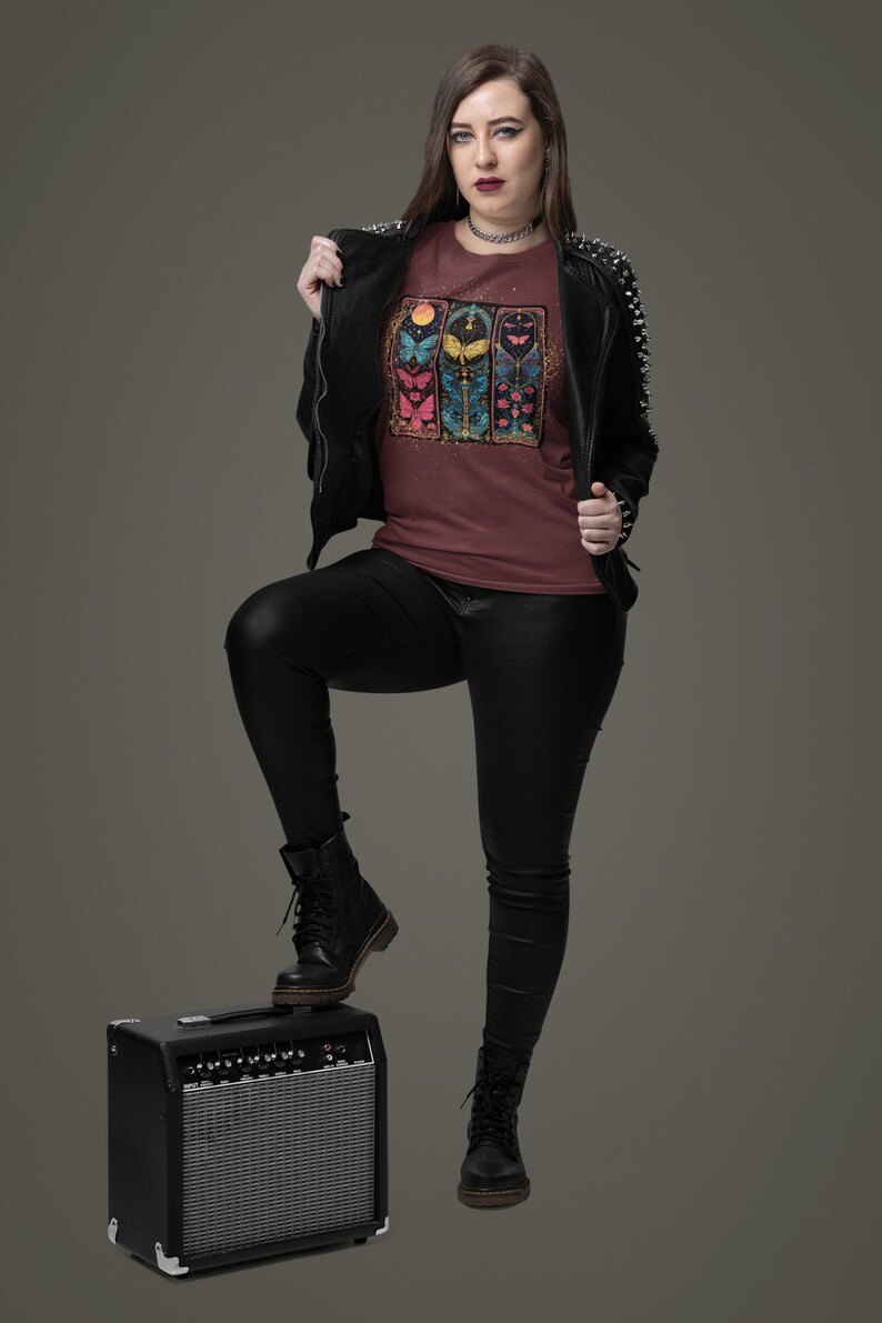 May include: A woman wearing a maroon t-shirt with a colourful butterfly graphic, a black leather jacket, black trousers, and black boots. She is standing with one foot on a black guitar amplifier.
