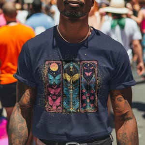 May include: A navy blue t-shirt featuring a graphic print of three vertical panels with butterflies, stars, and floral designs. Each panel has a gold border and a textured background.