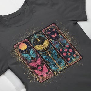 Könnte beinhalten: Ein dunkelgraues T-Shirt mit einem grafischen Druck von drei bunten Schmetterlings-Illustrationen in verzierten Rahmen. Die Schmetterlinge sind rosa, blau und gelb mit goldenen Akzenten. Der Hintergrund ist dunkelgrau mit einem dezenten gesprenkelten Muster.