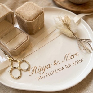 Könnte beinhalten: Eine weiße Keramik-Ringschale mit dem goldenen Text "Rüya & Mert" und "MUTLULUGA ILK ADIM". Die Schale enthält eine beige Samt-Doppelringschatulle, eine goldfarbene Schere und ein kleines, mit einem beigen Band gebundenes Trockenblumenarrangement.