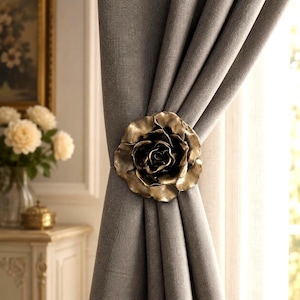 May include: A metallic gold-toned rose curtain tieback holding a thick, textured grey fabric curtain in place. The rose features layered, sculpted petals with a dark, antiqued finish in the center, creating a dimensional floral accent piece.