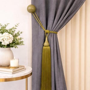 May include: A dark grey curtain held back by a decorative olive green tieback featuring a smooth spherical finial, a braided cord, and a long, flowing tassel. The curtain is draped vertically, showcasing its fabric texture and deep folds.