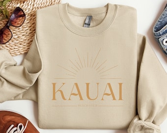Könnte beinhalten: Ein beiger Pullover mit einer goldenen Grafik einer Sonne mit Strahlen und dem Text "KAUAI BEACH CLUB".