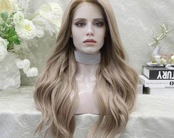 May include: A long, wavy blonde wig on a mannequin head. The wig has a natural-looking hairline and is styled with loose curls. The mannequin is wearing a silver rhinestone choker. The background includes white flowers and decorative items.