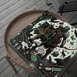 May include: A wrapped gift with a brown ribbon sits on a sheet of fantasy-themed wrapping paper. The paper features a green and brown illustration of an archer, forest elements, and dice. Scissors and tape are also visible.