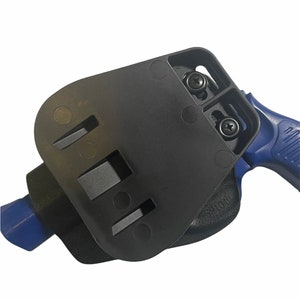 May include: Black polymer holster with a blue handgun. The holster features a rectangular design with two screws and several slots. The handgun's blue handle is visible. The holster is designed to securely hold a firearm.