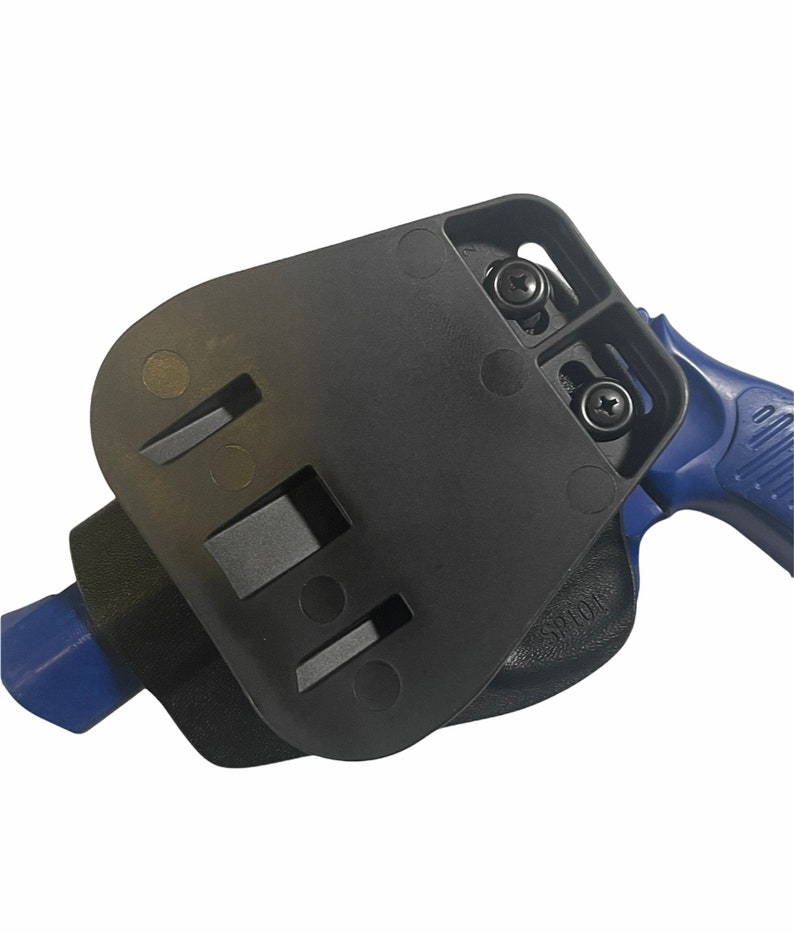 May include: Black polymer holster with a blue handgun. The holster features a rectangular design with two screws and several slots. The handgun's blue handle is visible. The holster is designed to securely hold a firearm.