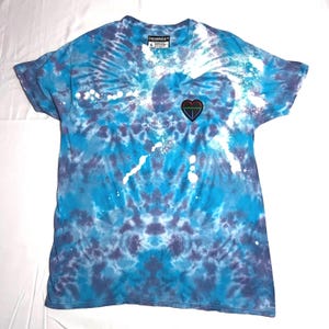 May include: A blue tie-dye t-shirt with a heart-shaped design and a small emblem. The shirt has a crew neck and short sleeves. The brand name "DEIBREST" is visible on the tag.