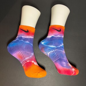 Puede incluir: Un par de calcetines teñidos con un borde y una puntera naranjas, que pasan al rojo, morado y azul. Un logotipo negro de Nike está en el borde. Los calcetines se muestran en pies de maniquí blancos.