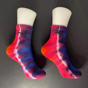 May include: A pair of colourful tie-dye ankle socks with a black Nike swoosh logo on each sock. The socks are a mix of red, orange, blue, and white.