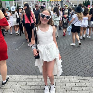 May include: A young person wearing a white dress with a high-low hemline and white trainers. They are wearing heart-shaped sunglasses and have a black bag slung over their shoulder. The person is standing on a brick path in front of a large building.