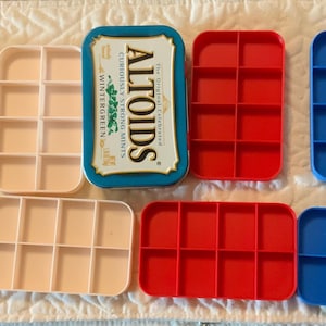 May include: A collection of rectangular plastic inserts with eight compartments each, arranged in black, light pink, red, and blue. One Altoids Wintergreen mint tin is positioned in the center, featuring a blue and gold label with white text.