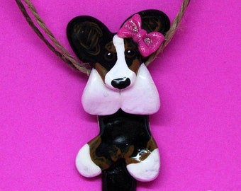 May include: A pendant shaped like a black, white, and brown dog with a pink glittery bow on its ear, suspended from a brown twine cord. The dog has a white chest, white paws, and a white tail tip. The text Paws in a Row Pet Art by Emily Rose is visible.