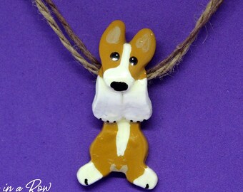 May include: A brown and white dog pendant suspended on a brown twine cord. The dog figure features upright ears, a white face stripe, and a black nose. The text Paws in a Row Pet Art by Emily Rose is printed in white in the bottom left corner.