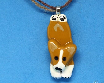 May include: A pendant shaped like a brown and white dog with a white blaze on its face, black eyes, and black paw pads, suspended from a brown twine cord. The text Paws in a Row Pet Art by Emily Rose is printed in the bottom right corner.
