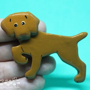 May include: A brown dog-shaped pin with a glossy finish, featuring a cartoon-style illustration of a dog with one front paw raised. The dog has black eyes, small black eyebrows, and a simple outline. The text Paws in a Row Pet Art by Emily Rose is visible.