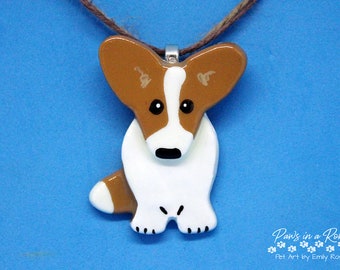 May include: A pendant shaped like a brown and white dog with large ears, black eyes, and a black nose, suspended from a brown cord. The text Paws in a Row Pet Art by Emily Rose is printed in white in the bottom right corner of the image.