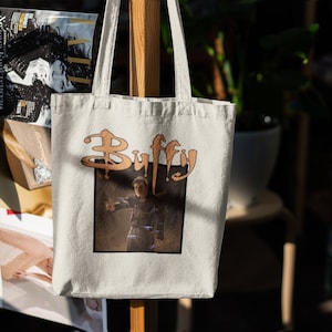 May include: A white canvas tote bag featuring a brown graphic of the word "Buffy" in a stylised font. The graphic also includes a picture of a person in a dark jacket standing in a forest.
