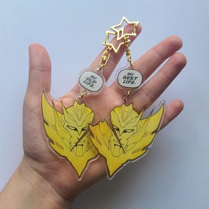 May include: Two yellow and clear acrylic keychains. Each keychain features a yellow character design with the text "MY OLD LIFE" and "MY BEST LIFE" in speech bubbles. The keychains are connected to a gold star-shaped charm.