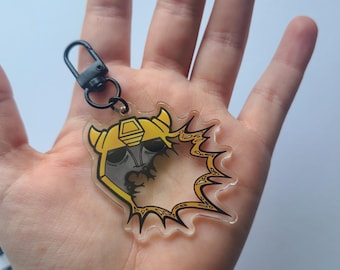 May include: A clear acrylic keychain featuring a yellow and gray cartoon character design with a black outline. The keychain has a black clasp and a circular burst design around the character's head.