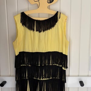 May include: A sleeveless yellow dress featuring multiple horizontal tiers of long black fringe. The fringe is layered across the chest and down the length of the garment, creating a distinct contrast against the bright yellow fabric base.