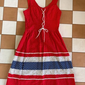 May include: A sleeveless red dress with white polka dots, featuring a white lace-up front closure with a bow. The skirt includes horizontal bands of white floral lace, a navy blue polka dot stripe, and white zigzag trim along the hem and waist.