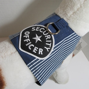 May include: A blue and white striped dog vest with a "SECURITY OFFICER" patch. The patch is black with white lettering and a white star. The vest has a silver metal ring attached.