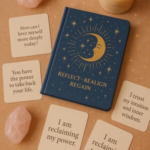 Könnte beinhalten: Ein marineblaues Notizbuch mit einem goldenen Halbmond- und Stern-Design, umgeben von Affirmationskarten und Rosenquarz-Kristallen. Das Notizbuch trägt die Aufschrift "REFLECT. REALIGN. REGAIN." Eine brennende Kerze steht daneben. Die Karten haben Affirmationen.