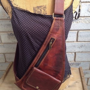 May include: A brown leather sling bag with a zipped main compartment and a front pocket. The bag features a long, adjustable strap for cross-body wear. The leather has a distressed finish, giving it a vintage look.