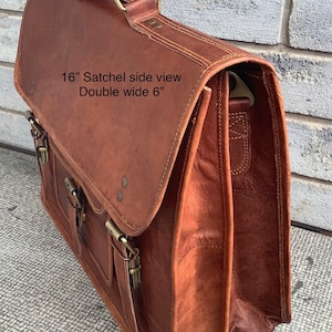 May include: A brown leather satchel with a double-wide design. The bag features a flap closure with two buckles and a top handle. The text on the bag reads "16" Satchel side view Double wide 6".