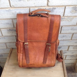 May include: A tan leather backpack with a flap closure, two front straps with buckles, and a top handle. The bag has a rectangular shape and appears to be made of distressed leather. The bag is sitting on a wooden surface.