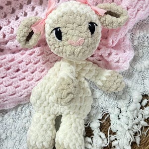 Pode incluir: Um brinquedo de pelúcia de cordeiro branco e bege de crochê com laços rosa. O cordeiro tem olhos pretos e um nariz rosa. O brinquedo está sentado em um cobertor branco e rosa.