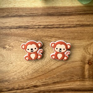 May include: Two small, brown monkey charms with white faces and white tails. The monkeys are sitting with their legs crossed and have a happy expression on their faces.