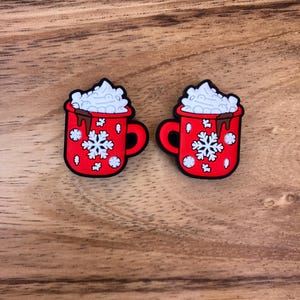 Peut inclure: Deux tasses en céramique rouge avec de la crème fouettée blanche et un filet de chocolat. Chaque tasse a un motif de flocon de neige et un bord blanc.