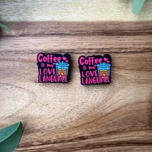Peut inclure: Deux breloques en caoutchouc noir avec le texte "Coffee is my love language" en lettres roses. Chaque breloque présente une image de dessin animé d'une tasse de café avec une paille.