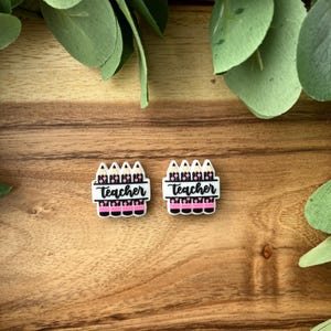 Peut inclure: Deux petits breloques en forme de crayon blanc avec le mot "Teacher" en script noir sur fond blanc, au-dessus d'un motif à rayures roses et noires. Les breloques sont sur une surface en bois, entourées de feuilles vertes.